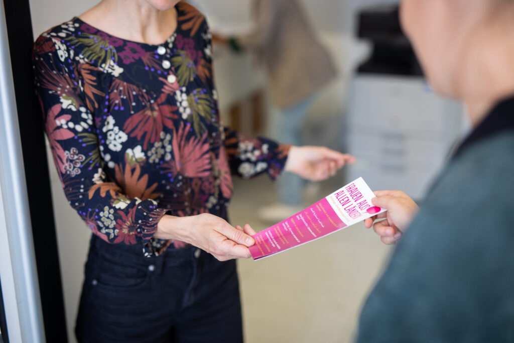 Zwei Personen, eine &uuml;bergibt der anderen einen pinken Flyer von Frauen aus allen L&auml;ndern mit Text